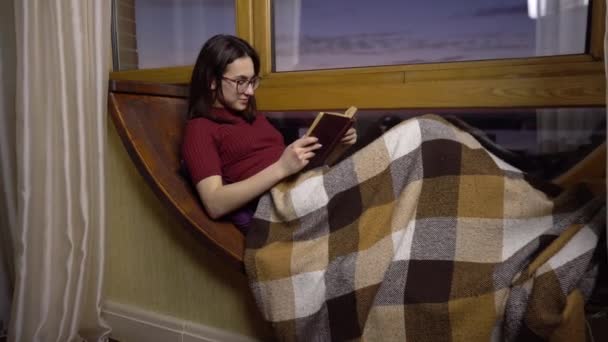 Una joven está leyendo un libro. Una chica yace en un alféizar junto a la ventana con un libro en las manos. Fuera de la noche — Vídeos de Stock