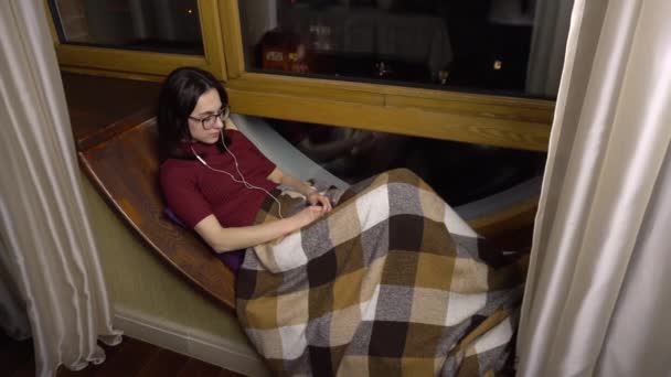 Een jonge vrouw luistert naar muziek op een koptelefoon. Een meisje ligt op een vensterbank bij het raam met een koptelefoon in haar oren. Buiten de avond. — Stockvideo