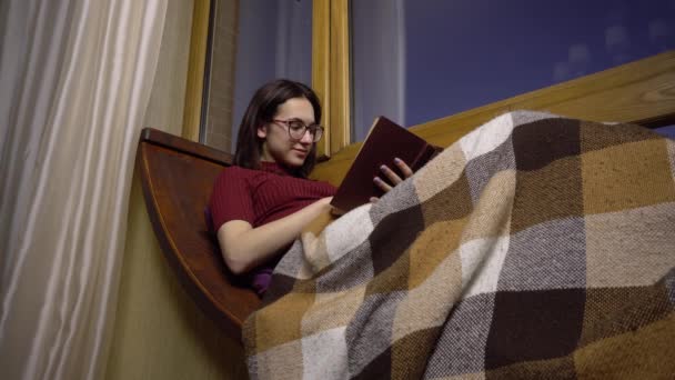 Μια νεαρή γυναίκα διαβάζει ένα βιβλίο. Ένα κορίτσι ξαπλώνει σε ένα περβάζι δίπλα στο παράθυρο με ένα βιβλίο στα χέρια της. Έξω από το βράδυ. — Αρχείο Βίντεο