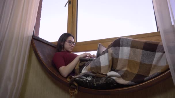 Una joven está escribiendo en un portátil. Una chica yace en un alféizar junto a la ventana con un portátil en sus manos. Fuera de la ventana un día brillante . — Vídeos de Stock