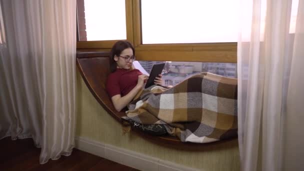 한 젊은 여성 이 태블릿으로 문자를 보내고 있습니다. 한 소녀 가손에 서판을 들고 창가 진열창에 누워 있다. 창문 밖은 화창 한 날. — 비디오
