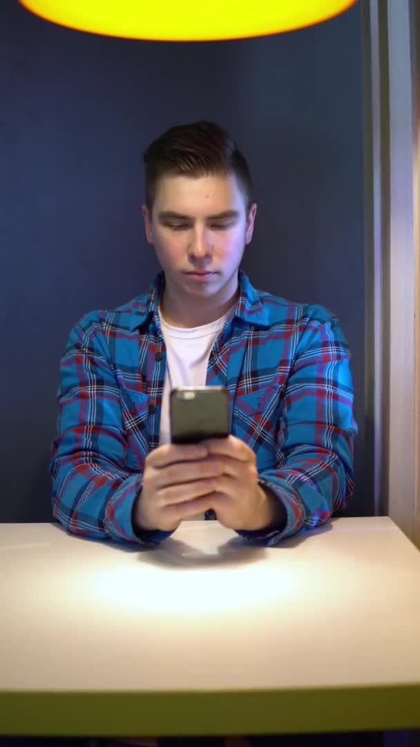 A young man with a phone sits at a table. A man sits at cafe at a table with a smartphone and is in correspondence. Vertical video. — Αρχείο Βίντεο