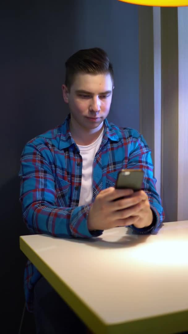 A young man with a phone sits at a table. A man sits at cafe at a table with a smartphone and is in correspondence. Vertical video. — Αρχείο Βίντεο