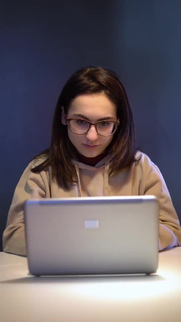 Una joven con un portátil se sienta en una mesa. Una chica está sentada en la cafetería en la mesa con un pequeño portátil y está escribiendo cartas. Vídeo vertical . — Vídeos de Stock