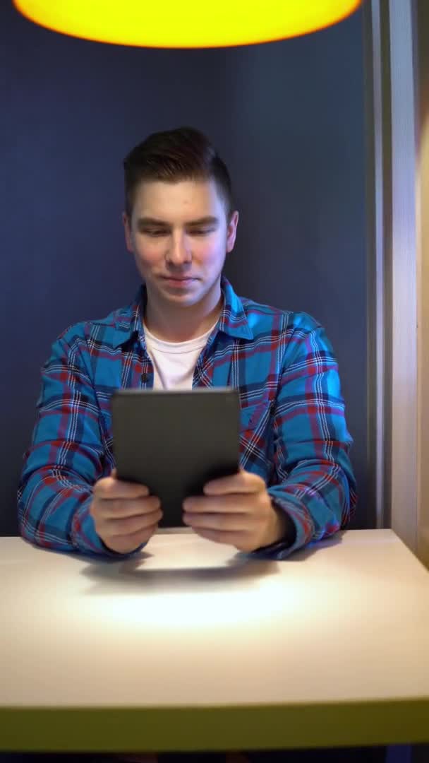 A young man with a tablet sits at a table. A man sits at home at a table with a tablet and is in correspondence. Vertical video. — Αρχείο Βίντεο