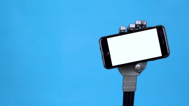 A mechanical hand holds a phone with a white screen. Gray cyborg hand holding a smartphone on a blue background. Template. — Stock Video
