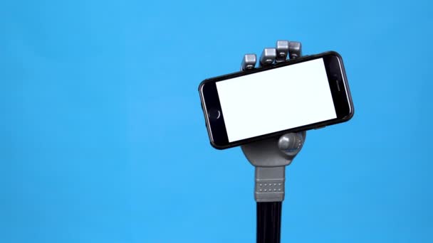 Een mechanische hand houdt een telefoon met een wit scherm vast. Grijze cyborg hand met een smartphone op een blauwe achtergrond. Template. — Stockvideo