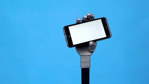 En mekanisk hand håller en telefon med en vit skärm. Grå cyborg hand håller en smartphone på en blå bakgrund. Mall. — Stockvideo