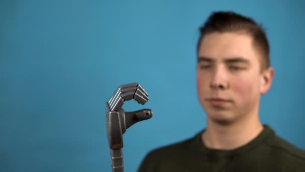 Młody człowiek patrzy na swoje mechaniczne ramię. Cyborg ściska palce na szarej dłoni na niebieskim tle. — Wideo stockowe