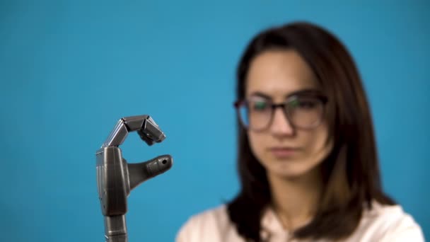 Een jonge vrouw kijkt naar haar mechanische arm. Cyborg knijpt vingers op een grijze hand op een blauwe achtergrond. — Stockvideo