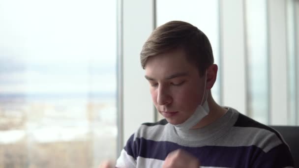 Een jongeman doet een medisch masker af, eet een hamburger en doet dan een masker op. Een man zit in een café bij het raam met een medisch masker op zijn gezicht. Preventie COVID-19. — Stockvideo