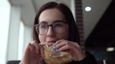 Genç bir kadın hamburger yiyor. Bir kız panoramik pencerenin yanındaki kafede oturur ve fast food yer..