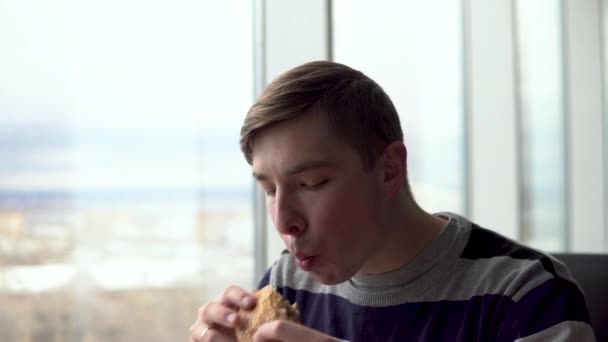 Молодой человек ест бургер. Мужчина сидит в кафе у панорамного окна и ест фаст-фуд . — стоковое видео