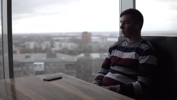 Un joven está escuchando música con auriculares. El hombre está sentado en un café junto a la ventana panorámica. Los auriculares están conectados al teléfono. Vista desde la ventana . — Vídeos de Stock