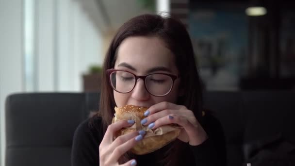 Egy fiatal nő hamburgert eszik. Egy lány ül egy kávézóban a panorámaablaknál, és gyorskaját eszik.. — Stock videók