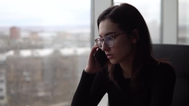 Una joven está hablando por teléfono. Una chica se sienta en un café junto a la ventana panorámica con un teléfono inteligente. Vista desde la ventana . — Vídeos de Stock