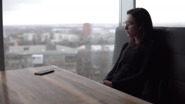 Una joven está escuchando música con auriculares. La chica está sentada en un café junto a la ventana panorámica. Los auriculares están conectados al teléfono. Vista desde la ventana . — Vídeos de Stock