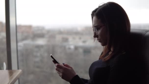 Egy fiatal nő telefonnal ül az asztalnál. Egy lány ül egy kávézóban a panorámaablaknál okostelefonnal a kezében. Kilátás az ablakból. — Stock videók
