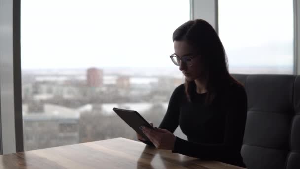 En ung kvinna med en tablett sitter vid ett bord. En flicka sitter på ett café vid panoramafönstret med en surfplatta i händerna. Utsikt från fönstret. — Stockvideo