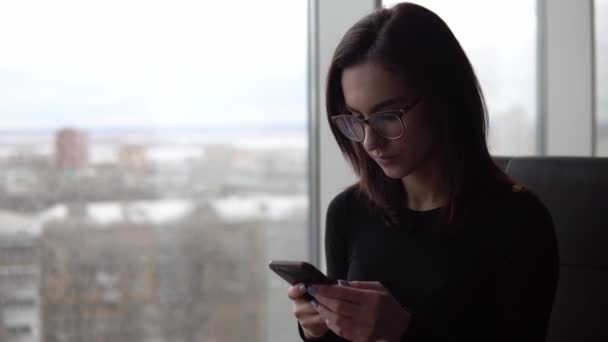 Una joven con un teléfono. Una chica se sienta en un café junto a la ventana panorámica con un teléfono inteligente en sus manos. Vista desde la ventana . — Vídeos de Stock