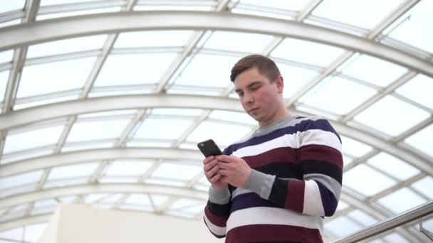 Młody człowiek stoi z telefonem. Mężczyzna używa smartfona przeciwko szklanemu dachowi. Zajęta osoba pisze do posłańca.. — Wideo stockowe