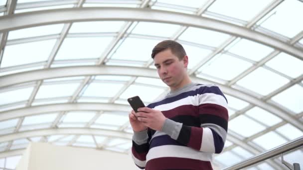 Ένας νεαρός άνδρας στέκεται με ένα τηλέφωνο. Ένας άντρας χρησιμοποιεί ένα smartphone ενάντια σε μια γυάλινη οροφή. Ένας πολυάσχολος άνθρωπος στέλνει μήνυμα στον αγγελιοφόρο.. — Αρχείο Βίντεο