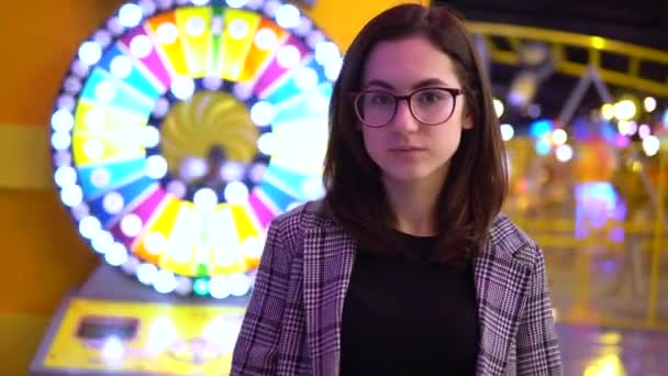 Una joven está parada en un parque de diversiones. La cámara se acerca a la chica. Parque para niños con máquinas tragamonedas y bombillas brillantes . — Vídeo de stock