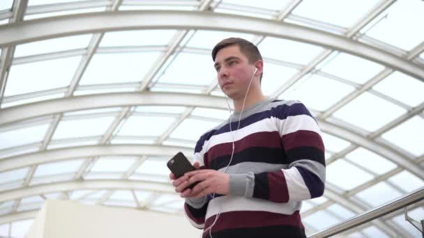 Een jongeman staat in een koptelefoon. Een man luistert naar muziek op een koptelefoon tegen een dak van een pan-glas. — Stockvideo