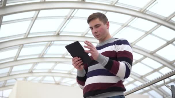 Un giovane uomo è in piedi con una tavoletta. Un uomo usa una tavoletta contro un tetto di vetro. Una persona impegnata sta messaggiando nel messaggero . — Video Stock
