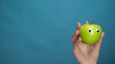 Kadının elinde gözleri olan yeşil bir elma. Apple çerçevenin içine atlar ve mavi bir arkaplanda etrafına bakar. Kadın eli yakın çekim.