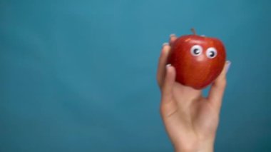 Kadın elinde gözleri olan kırmızı bir elma. Apple çerçevenin içine atlar ve mavi bir arkaplanda etrafına bakar. Kadın eli yakın çekim.