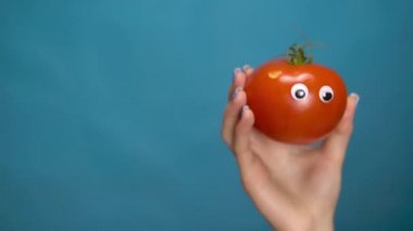 Kadının elinde gözleri olan kırmızı domates. Domates çerçevenin içine atlar ve mavi bir arkaplanda etrafına bakar. Kadın eli yakın çekim.