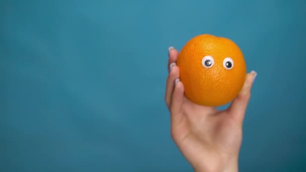 女性の手に目でオレンジ。オレンジがフレームに飛び込み、青の背景を見回します。女の手をクローズアップ. — ストック動画