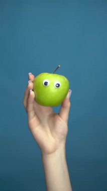 Bir kadının elinde gözleri olan yeşil elma. Yaklaş. Apple mavi bir arka planda etrafına bakıyor. Dikey video.