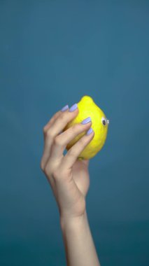 Bir kadının elinde gözleri olan limon. Yakın plan. Lemon mavi bir arka planda etrafına bakıyor. Dikey video.