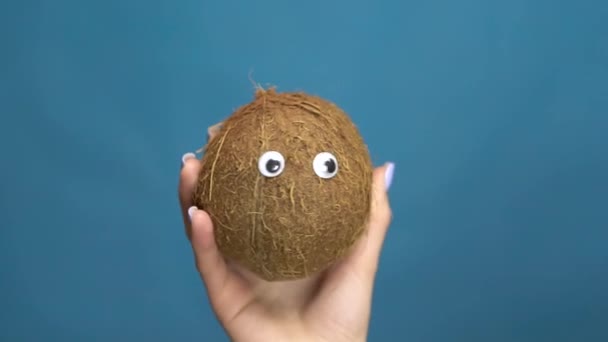 Coconut with eyes in a woman hand close-up. Coconut shakes and twists eyes on a blue background. Slow motion — Stock Video