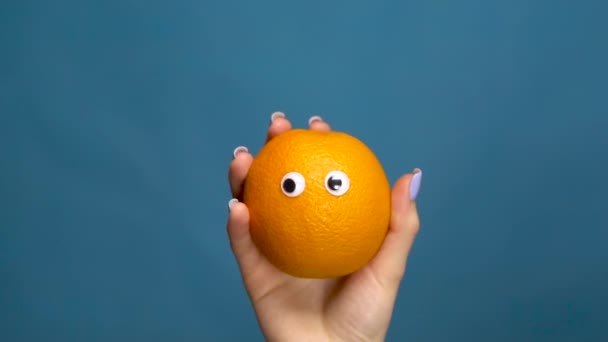 Laranja com olhos em uma mão mulher close-up. Laranja treme e torce os olhos em um fundo azul. Movimento lento . — Vídeo de Stock