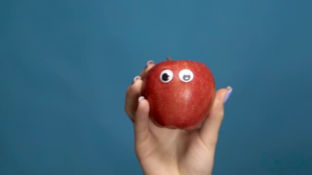 Manzana roja con ojos en una mano de mujer de cerca. Apple tiembla y tuerce los ojos sobre un fondo azul. Movimiento lento . — Vídeo de stock