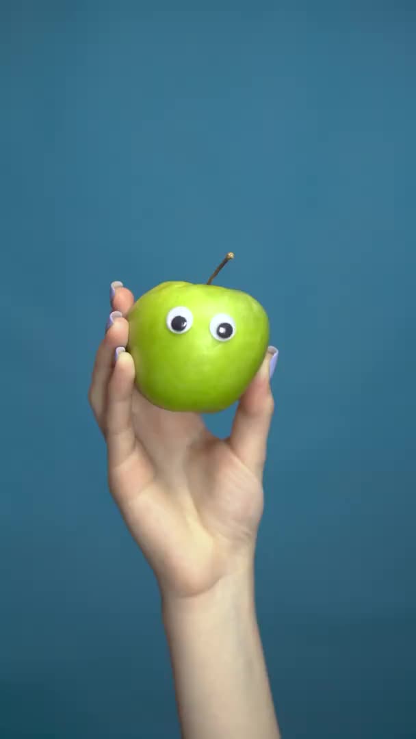Manzana verde con ojos en una mano de mujer de cerca. Apple mira a su alrededor sobre un fondo azul. Vídeo vertical . — Vídeo de stock