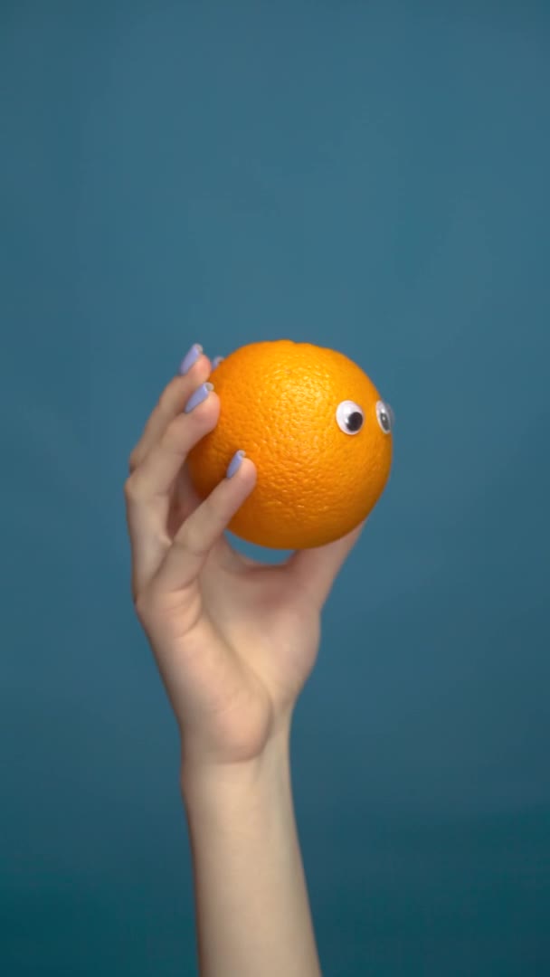 Orange mit Augen in einer Frauenhand aus nächster Nähe. Orange sieht sich auf blauem Hintergrund um. Vertikales Video. — Stockvideo