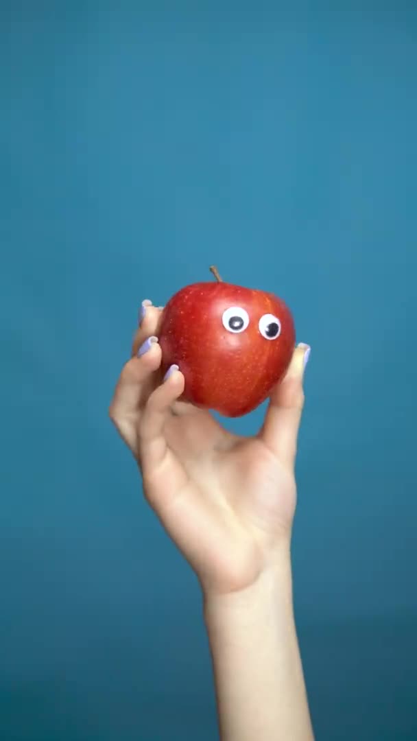 Manzana roja con ojos en una mano de mujer de cerca. Apple mira a su alrededor sobre un fondo azul. Vídeo vertical . — Vídeos de Stock