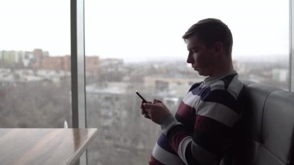 Un joven con un teléfono se sienta en una mesa. Un hombre se sienta en un café junto a la ventana panorámica con un teléfono inteligente en sus manos. Vista desde la ventana . — Vídeo de stock