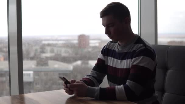 Un joven con un teléfono se sienta en una mesa. Un hombre se sienta en un café junto a la ventana panorámica con un teléfono inteligente en sus manos. Vista desde la ventana . — Vídeos de Stock