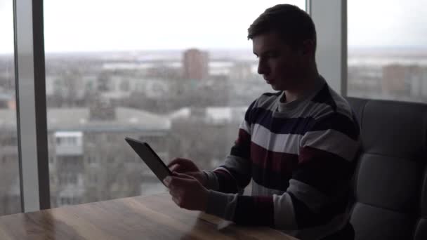 Un joven con una tablilla se sienta a la mesa. Un hombre se sienta en un café junto a la ventana panorámica con una tableta en las manos. Vista desde la ventana . — Vídeos de Stock