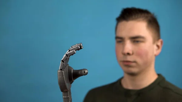 Un joven mira su brazo mecánico. Brazo Cyborg sobre una mano gris sobre un fondo azul . — Foto de Stock