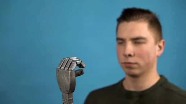 Młody człowiek patrzy na swoje mechaniczne ramię. Cyborg ramię na szarej dłoni na niebieskim tle. — Zdjęcie stockowe
