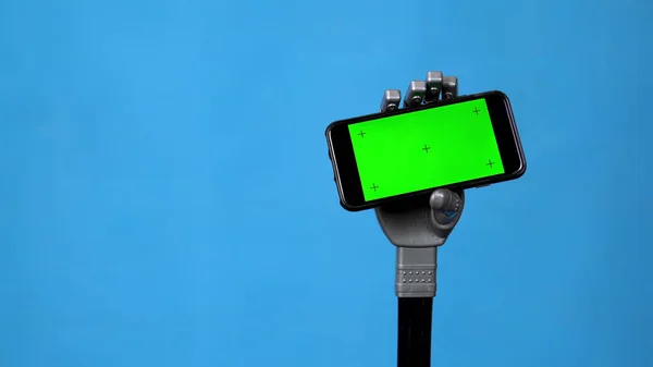 Механічна рука тримає телефон із зеленим екраном. Сіра рука кіборга тримає смартфон на синьому фоні. Клавіша Chroma . — стокове фото