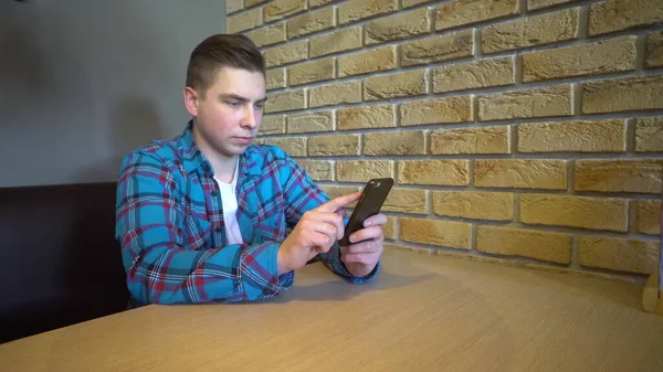 Um jovem com um telefone senta-se à mesa. Um homem senta-se em casa em uma mesa com um telefone e está em correspondência . — Fotografia de Stock