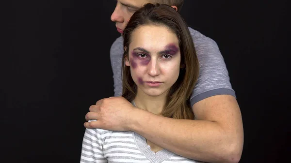 A young man hugs a young woman. A woman with bruises on her face. Quarrel in a young family. Domestic violence. On a black background — Stock Photo, Image