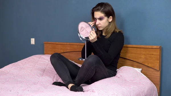 A young woman examines bruises on her face in the mirror. Quarrels in a young family. Domestic violence. The girl is sitting on the bed — Stock Photo, Image